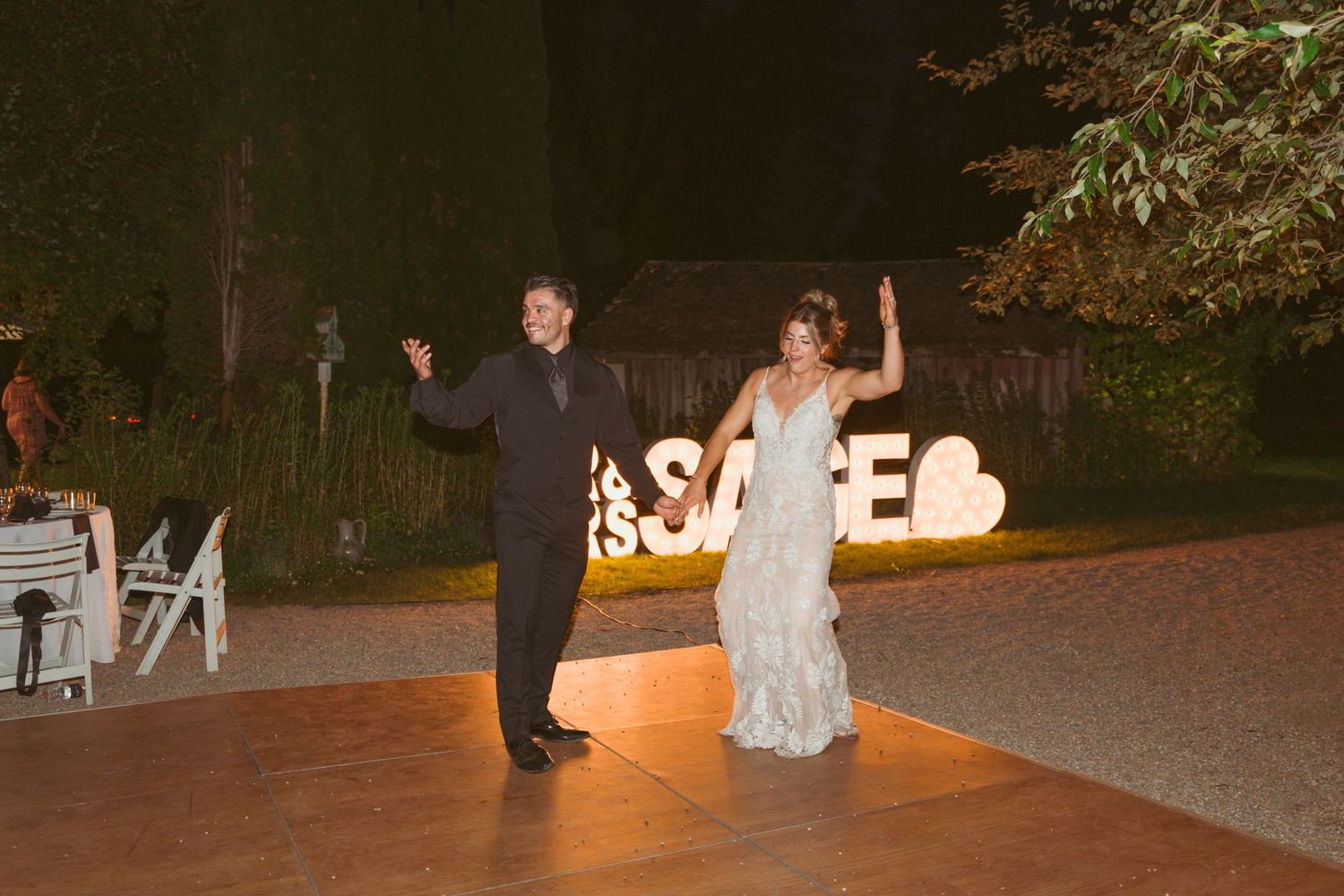 Couple celebrating with raised arms in front of illuminated sign at outdoor wedding reception.