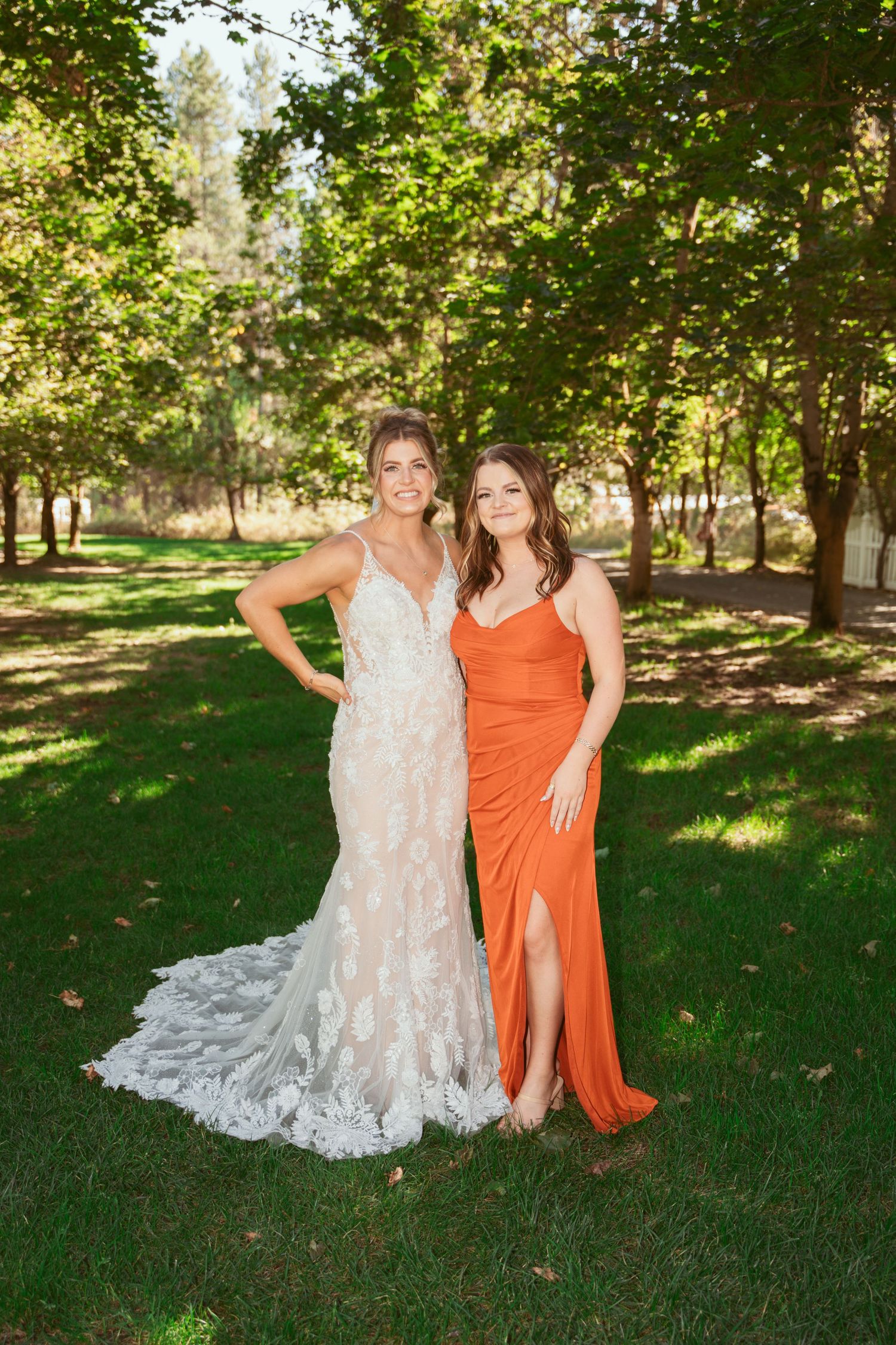 Wedding party members posing together on grass under trees wearing white and orange dresses.