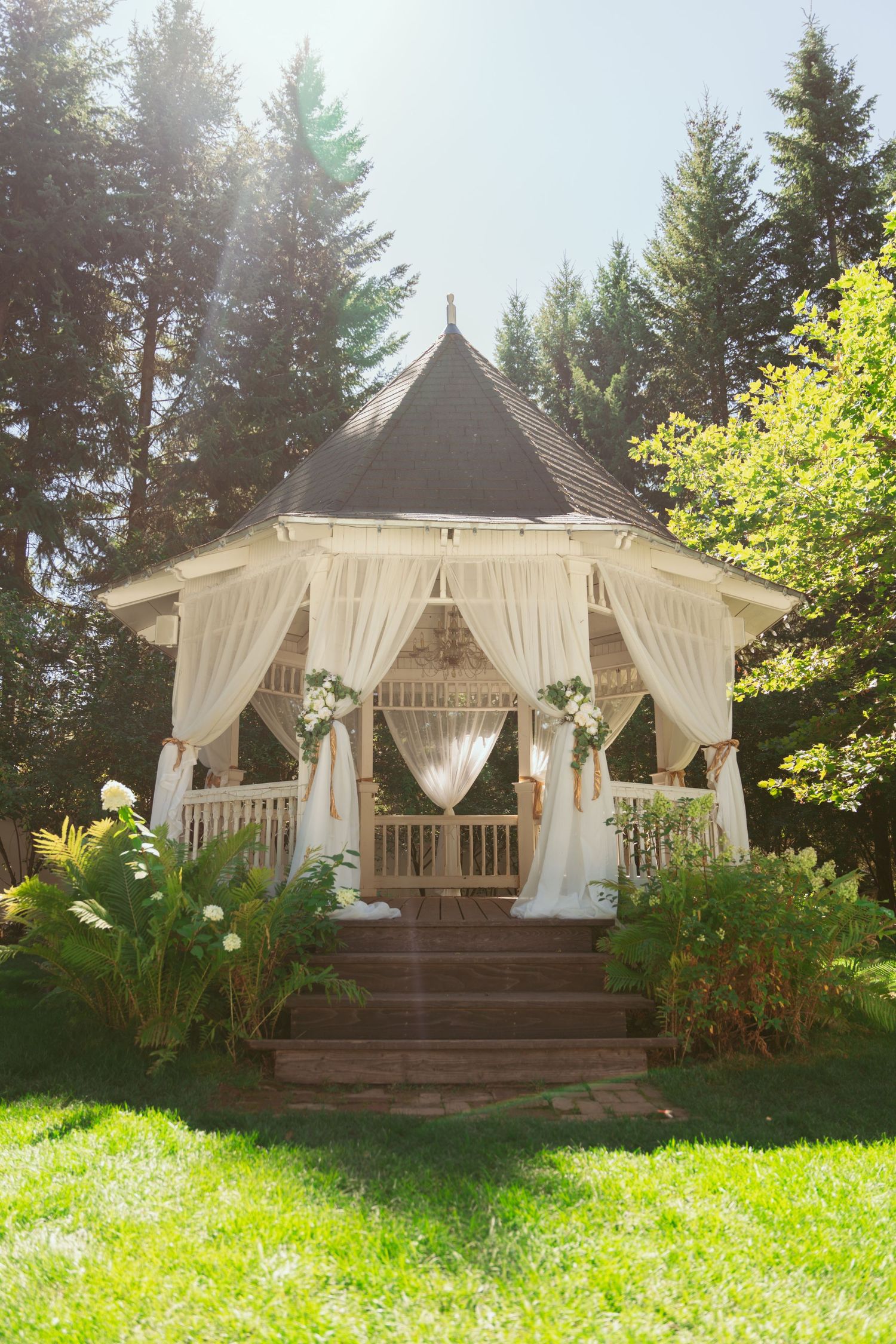 A white gazebo decorated with flowing curtains and floral arrangements sits in a sunlit garden setting.