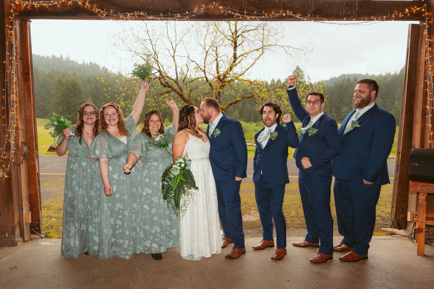 Wedding party celebrates joyfully under wooden arch with natural landscape behind them.