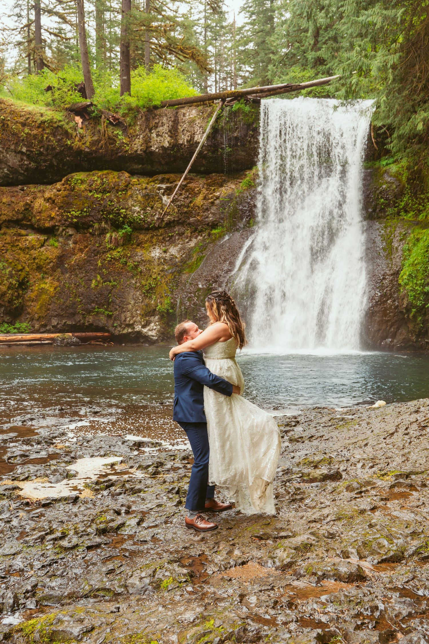 Wedding couple dances on rocky ground with majestic waterfall backdrop.