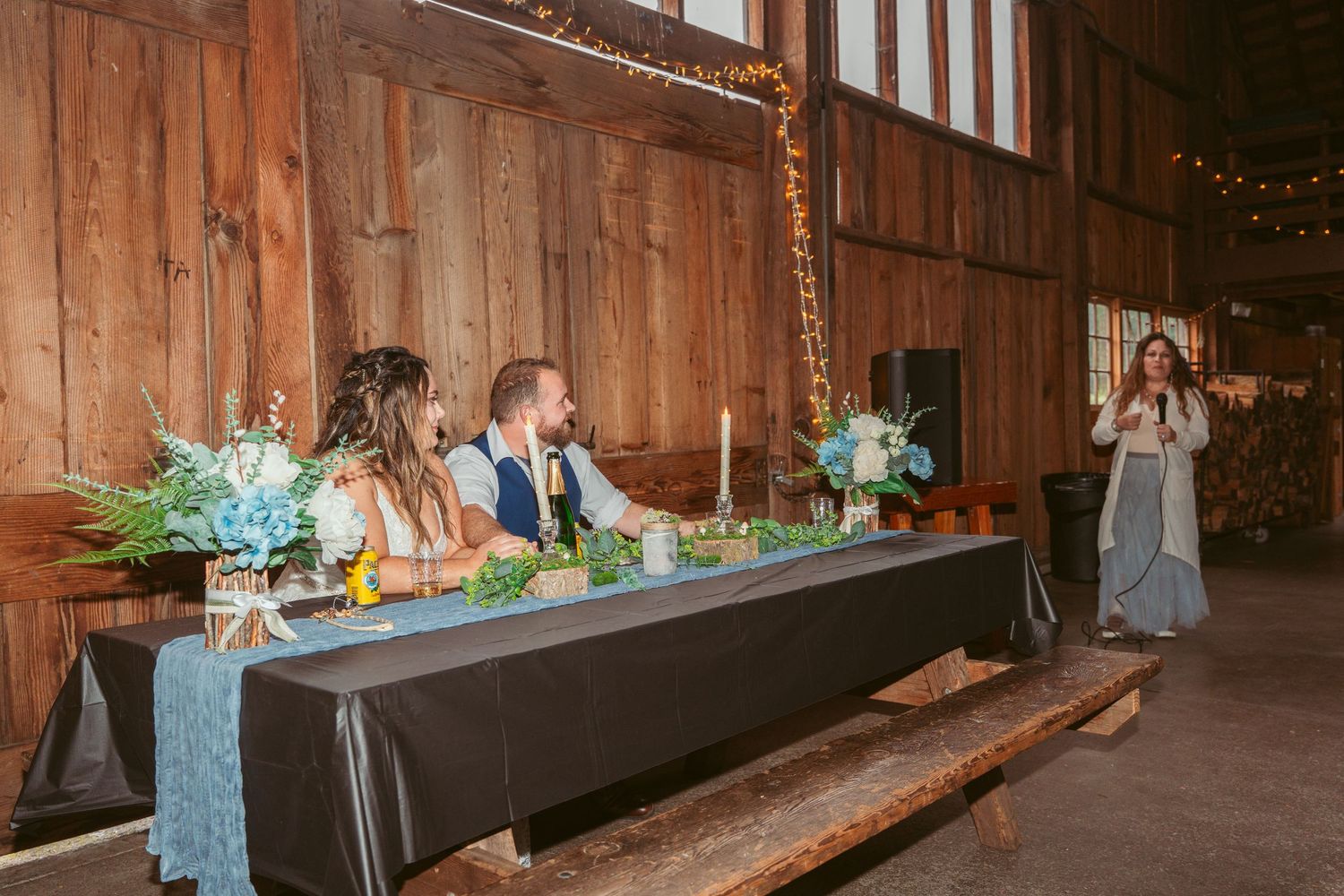 Long wooden benches line the interior of a barn decorated with string lights and floral arrangements.