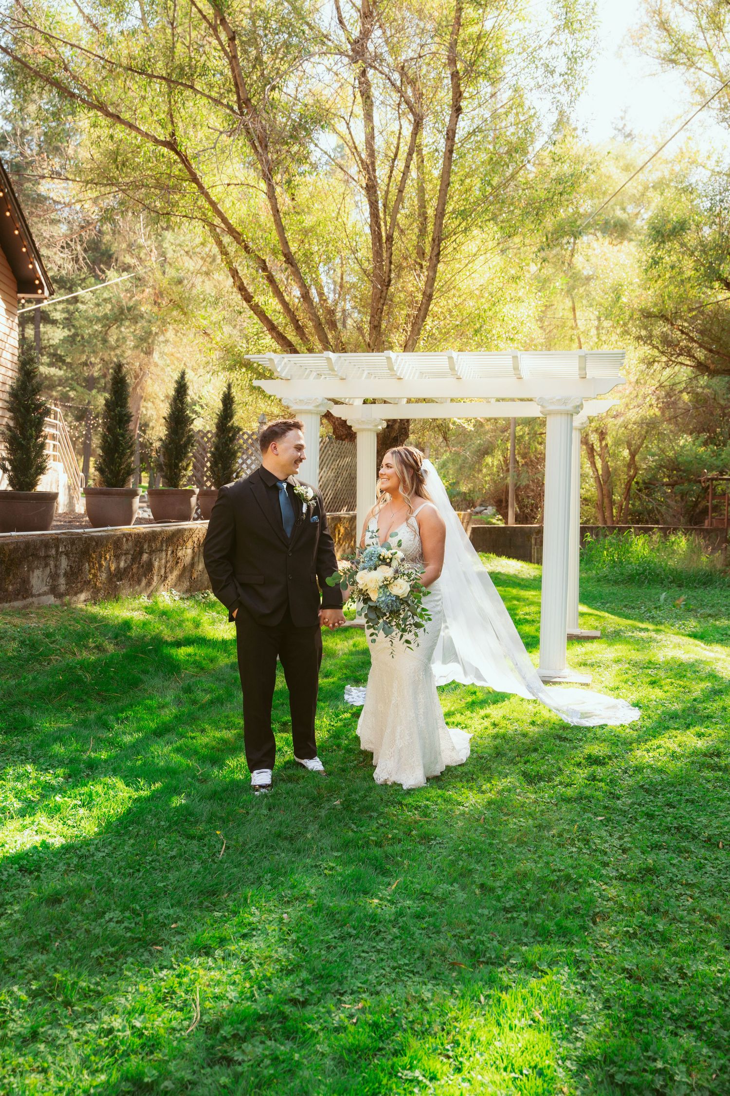 Couple shares a romantic moment under a white pergola surrounded by lush greenery and trees in an outdoor setting.