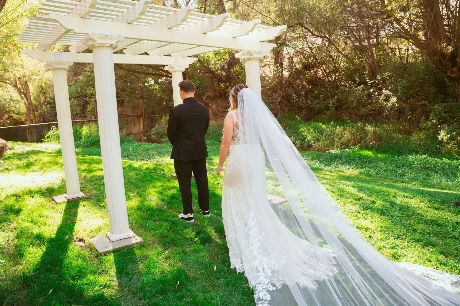A wedding couple stands under a white pergola in a sunlit garden setting with a flowing cathedral veil.