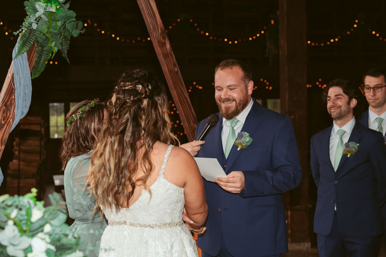 Wedding party gathers in rustic barn venue with decorative lighting and greenery.