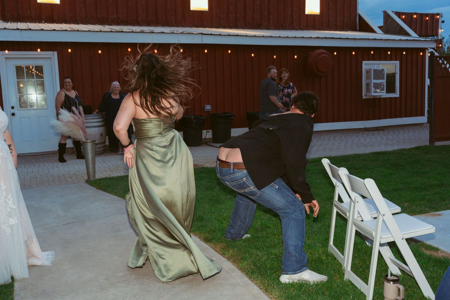 Wedding party members dance and celebrate on a lawn with white folding chairs in the background.