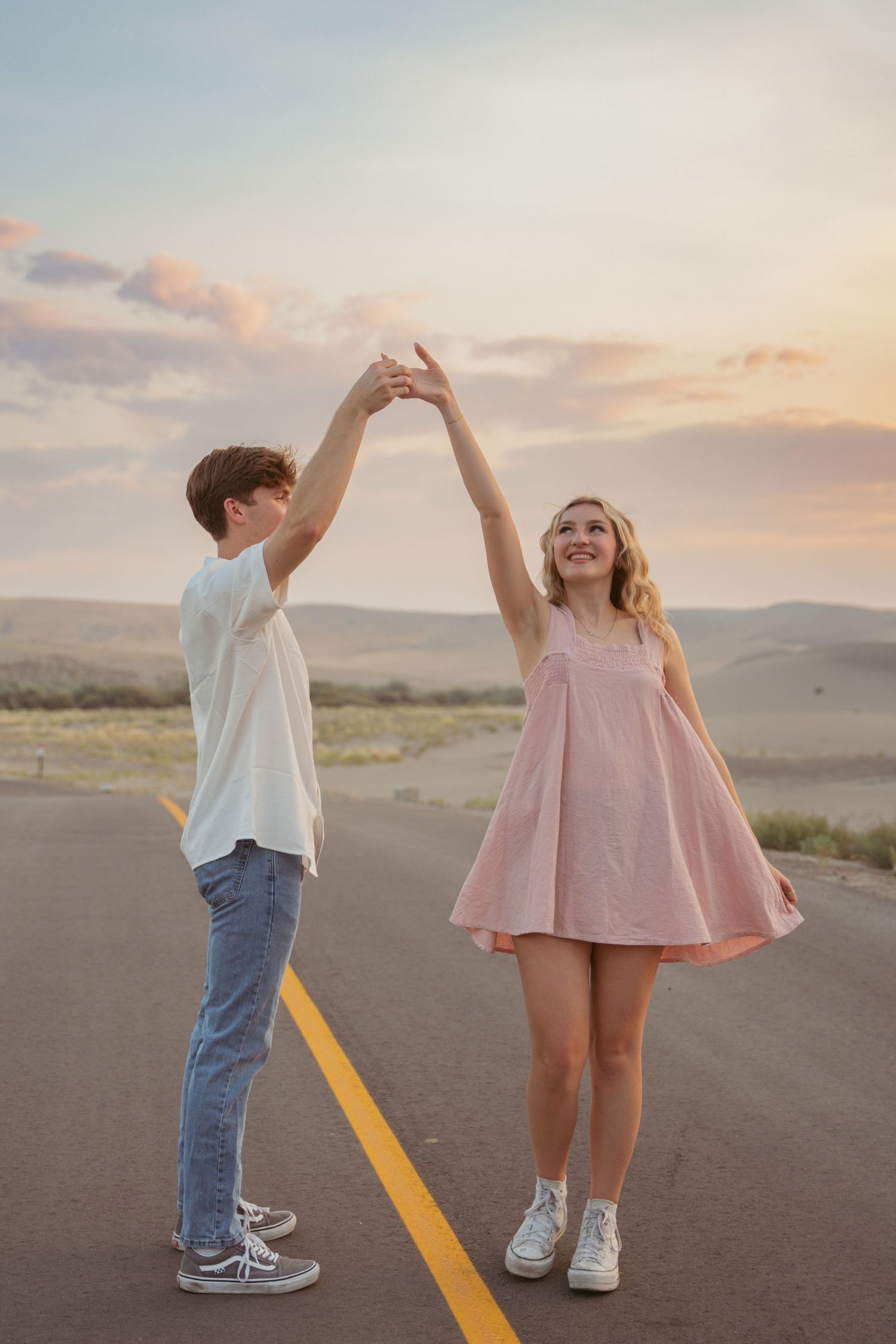 A couple holds hands while dancing on a rural road at sunset.