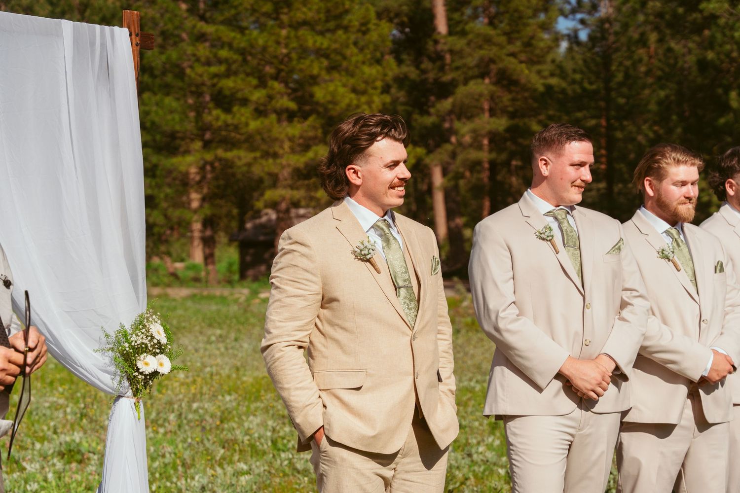 Groomsmen in beige suits stand at attention during outdoor forest wedding ceremony.