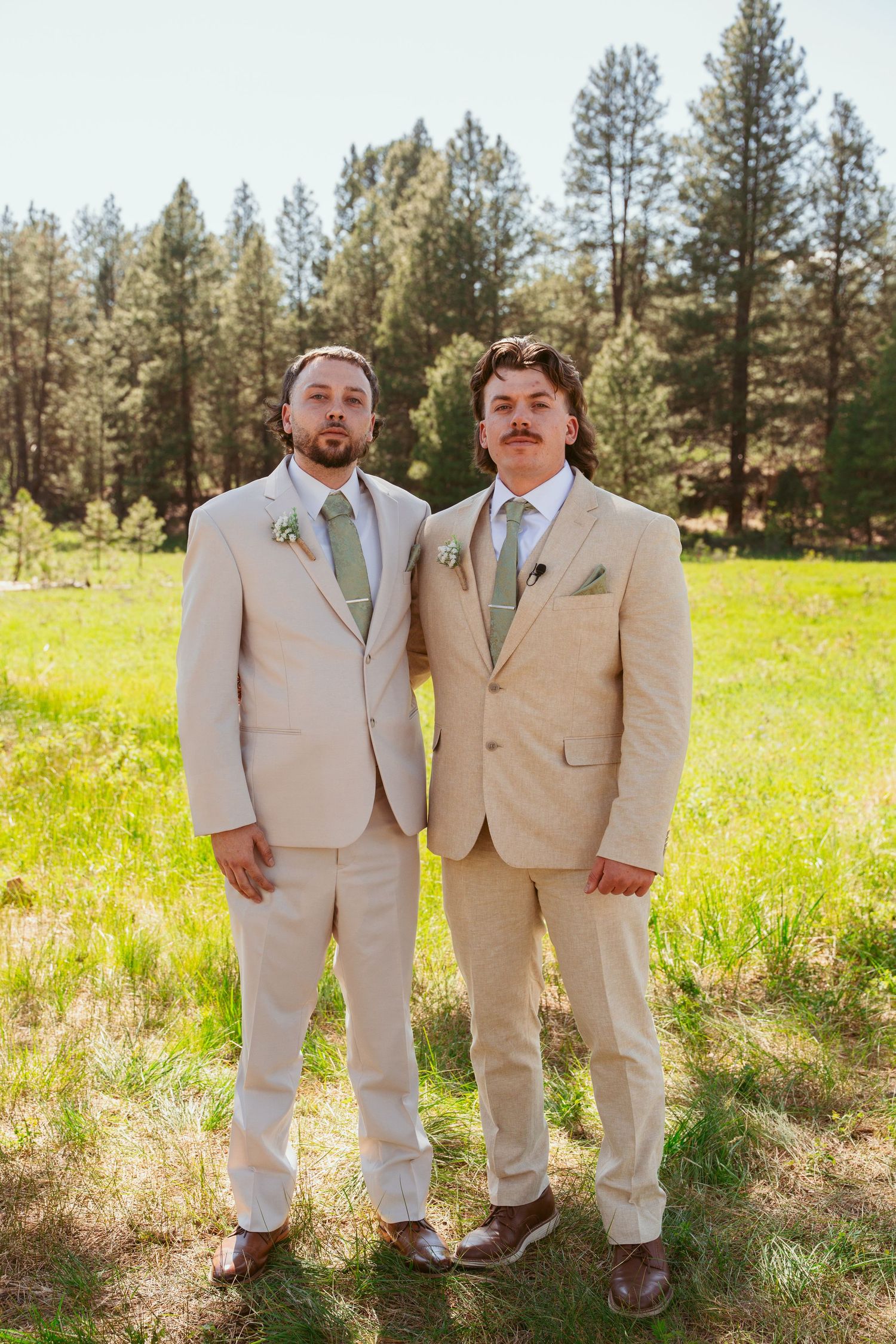 Two groomsmen in beige suits pose playfully in a sunny meadow with pine trees in the background.
