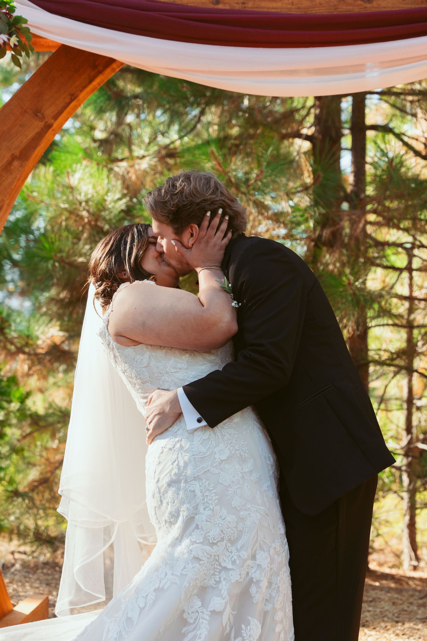 A romantic first kiss as newlyweds under a draped white fabric canopy in a forest setting.