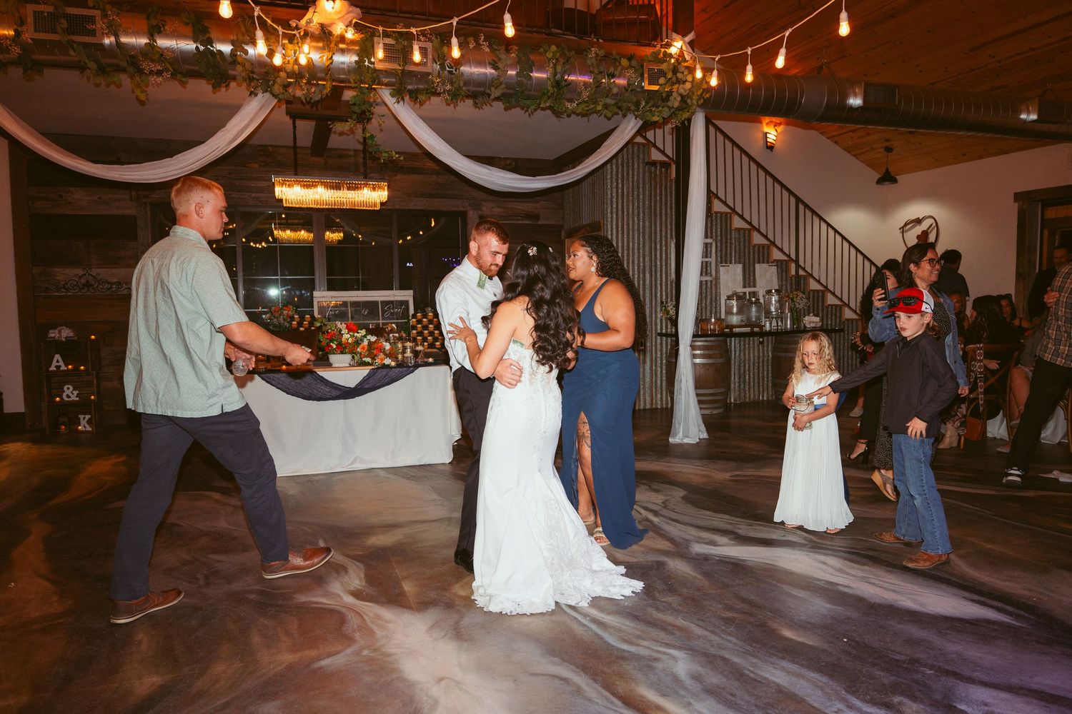 Wedding guests dance and celebrate on a rustic venue dance floor under warm string lights.