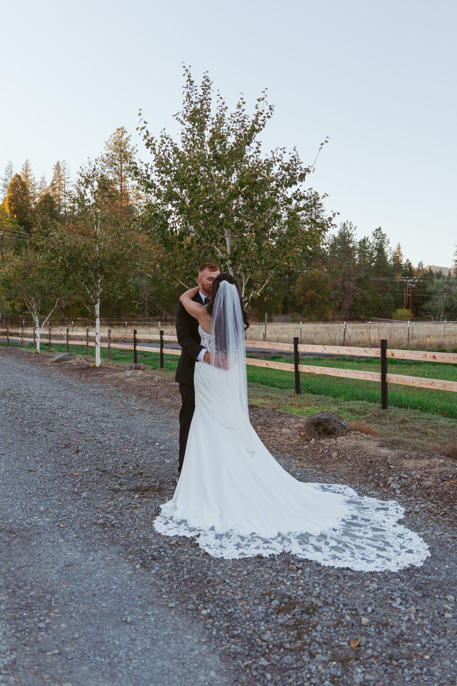 Long lace wedding train trails beautifully across gravel path during outdoor photo session.