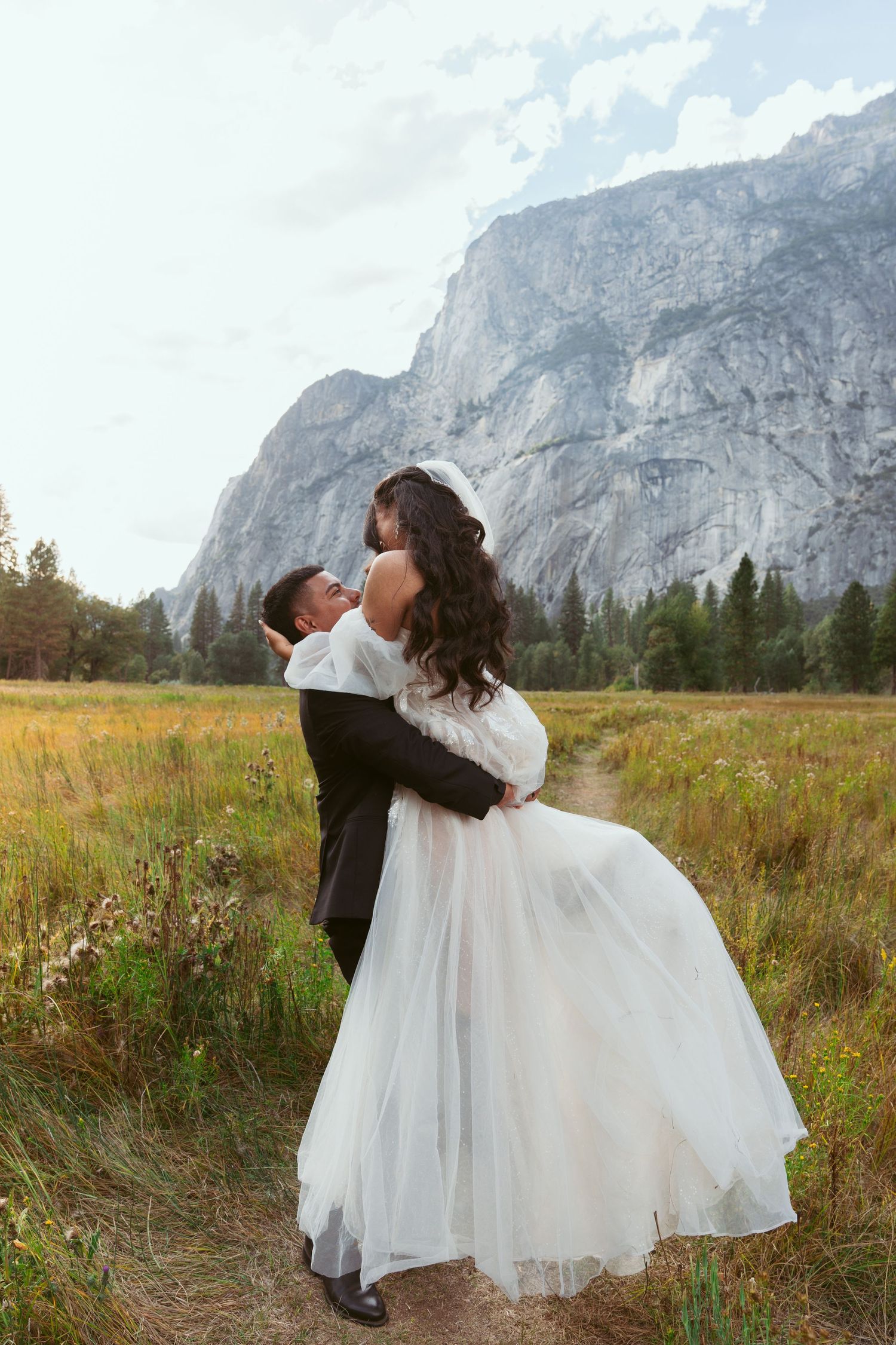 Dancing couple in wedding attire spins through wildflower meadow beneath Yosemite cliffs.
