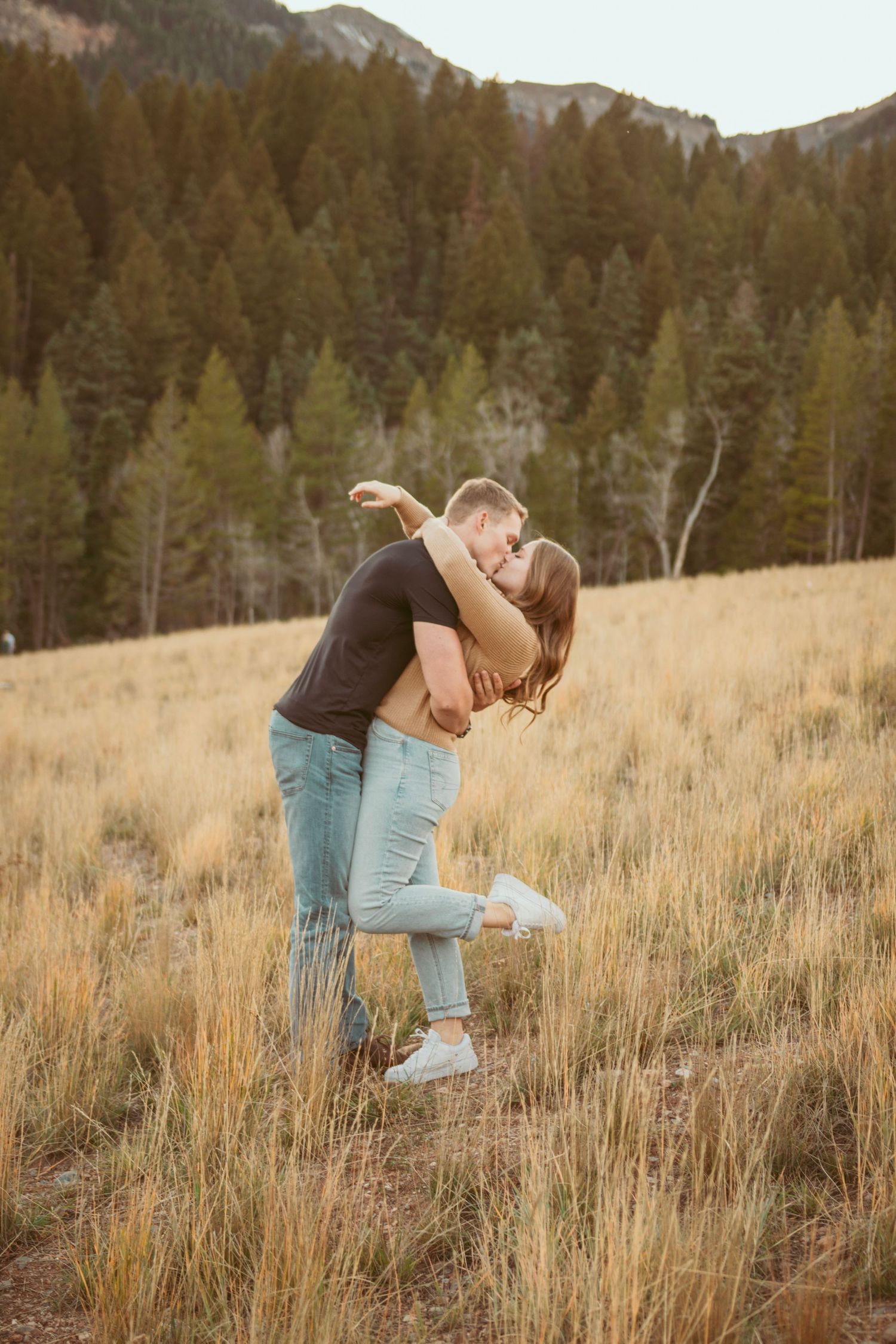 A couple embraces in a mountain meadow with evergreen forests in the background.