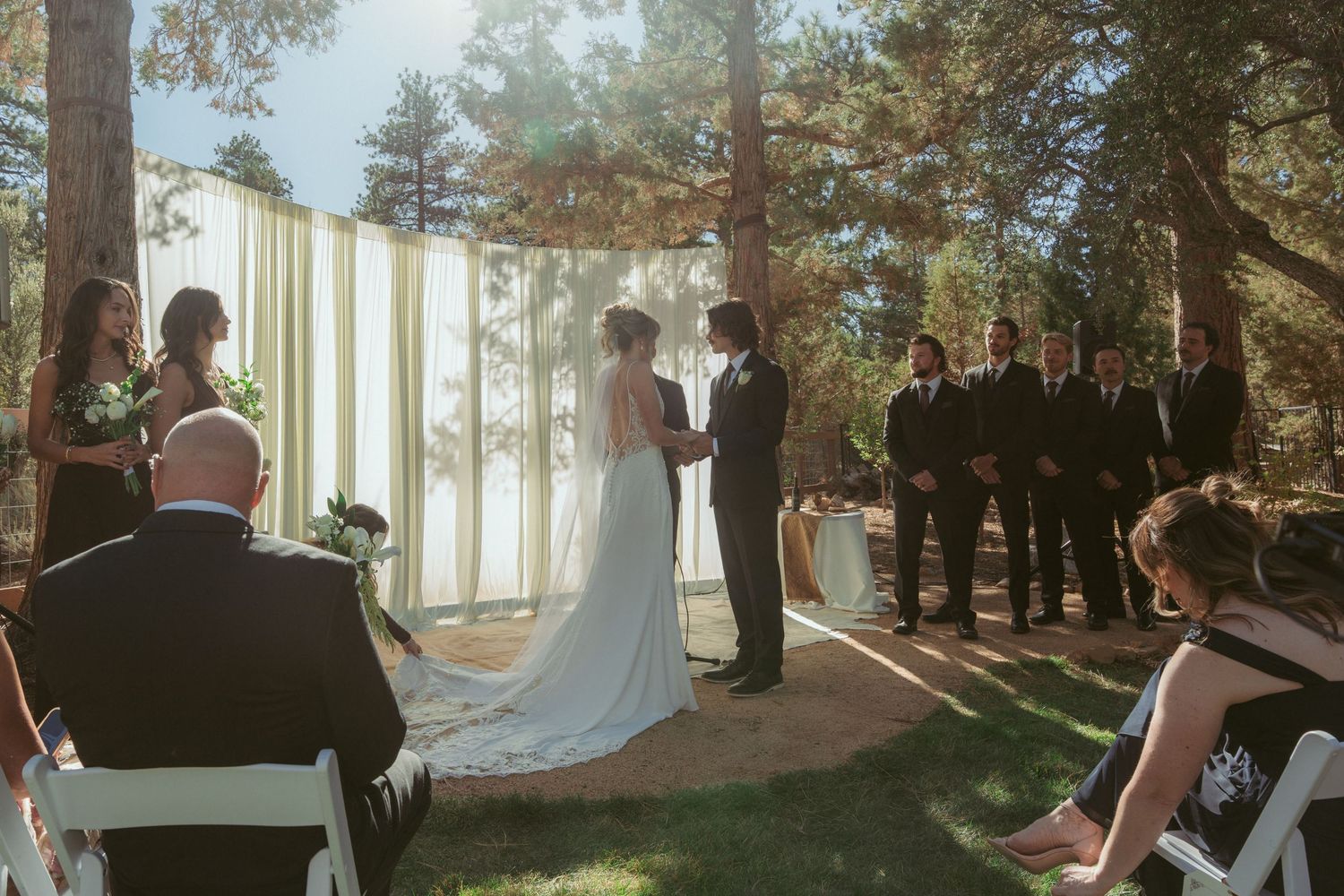 An outdoor wedding ceremony in a forest setting with white curtains and wooden chairs.