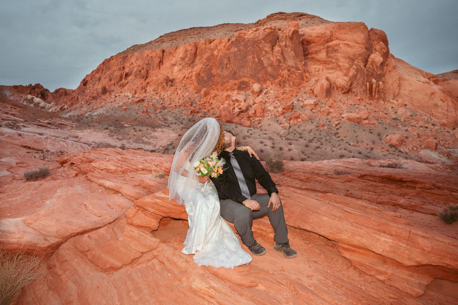 Wedding couple poses dramatically on red rock formations in a desert landscape.