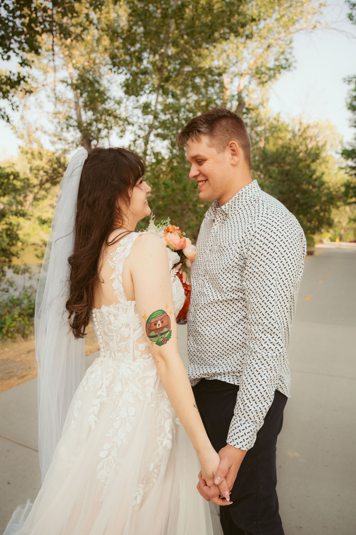 A couple in wedding attire embraces while holding hands on a sunlit path.