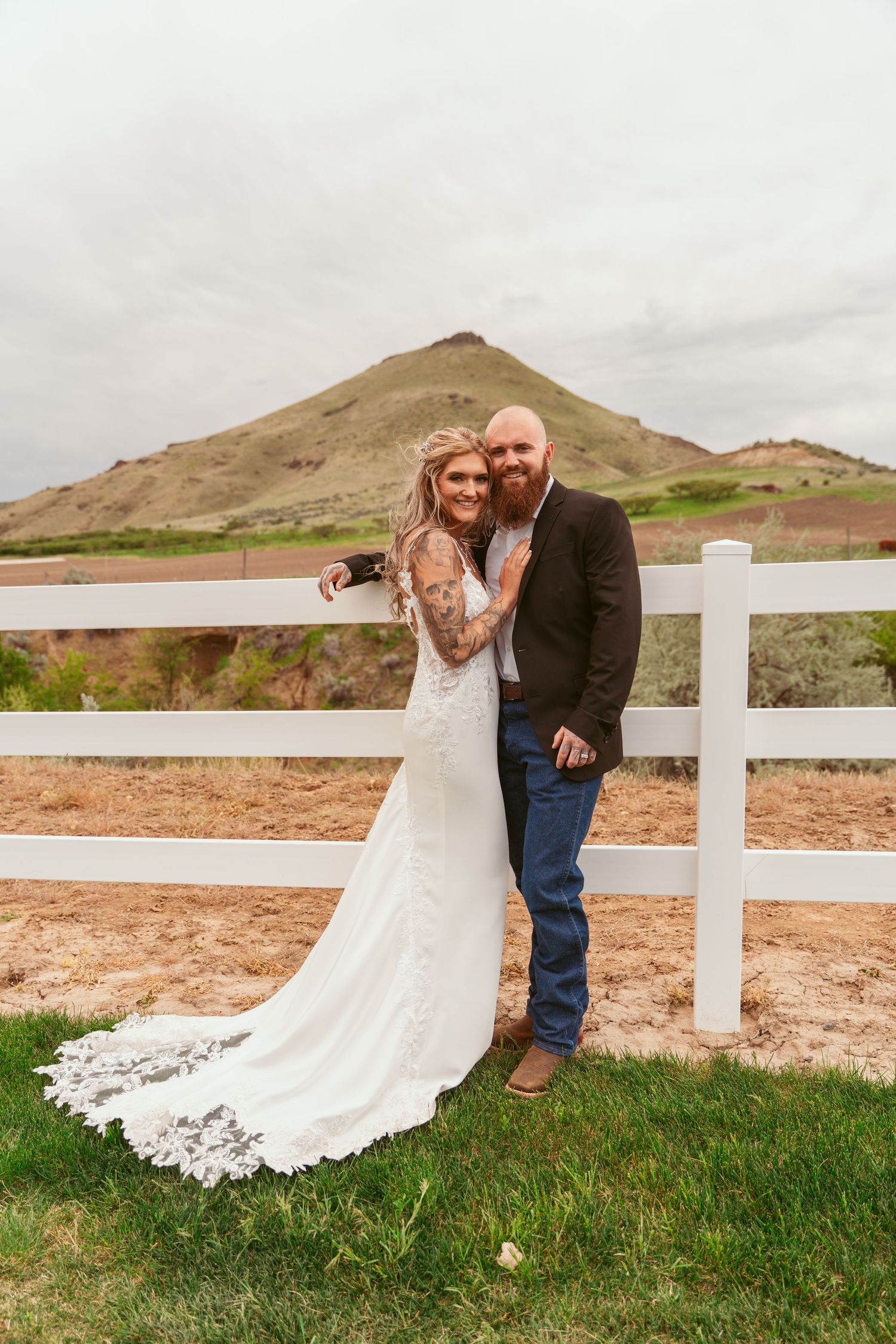 Newlyweds standing by white fence with mountain backdrop on their wedding day.