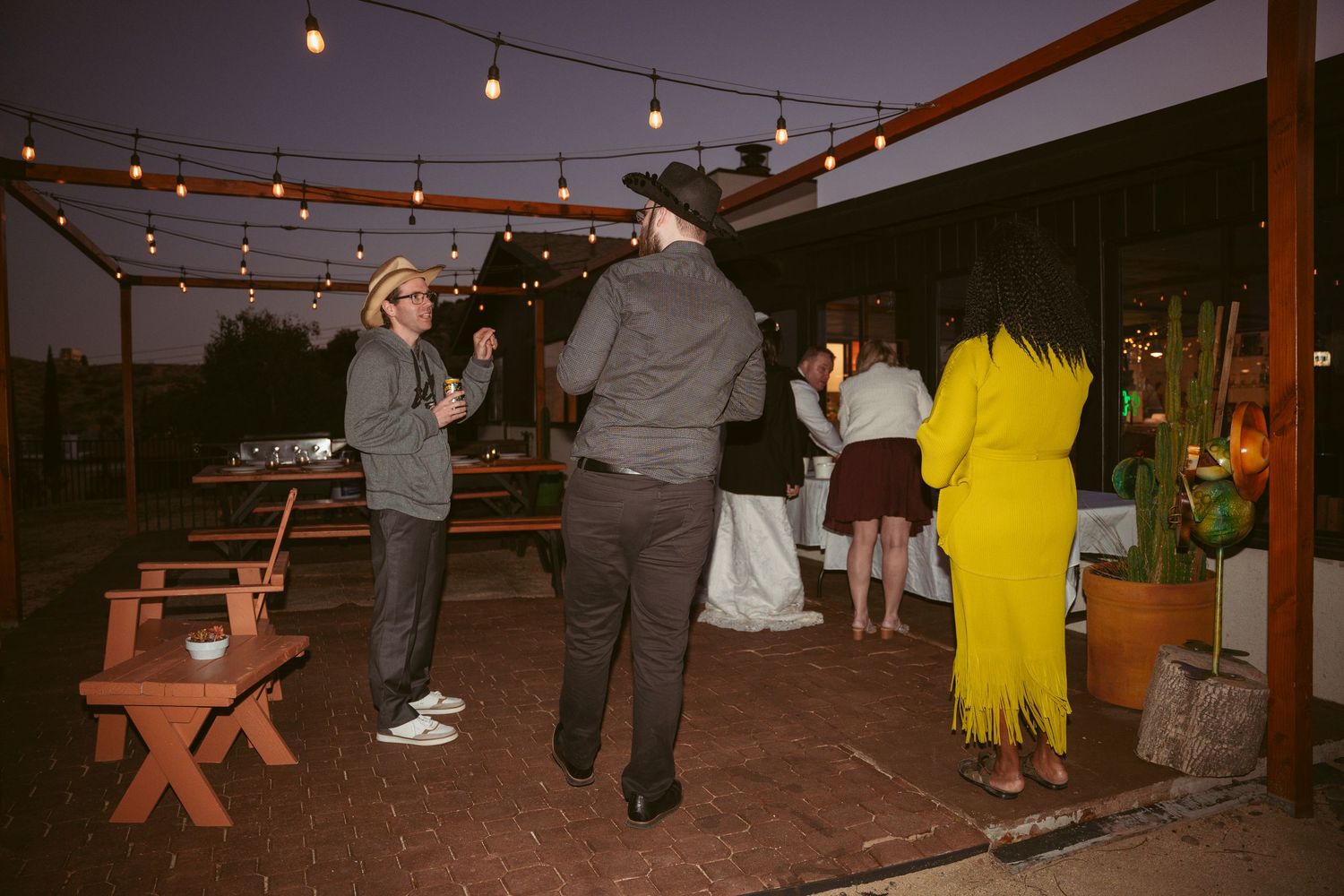 Outdoor reception area decorated with hanging string lights and wooden picnic tables.