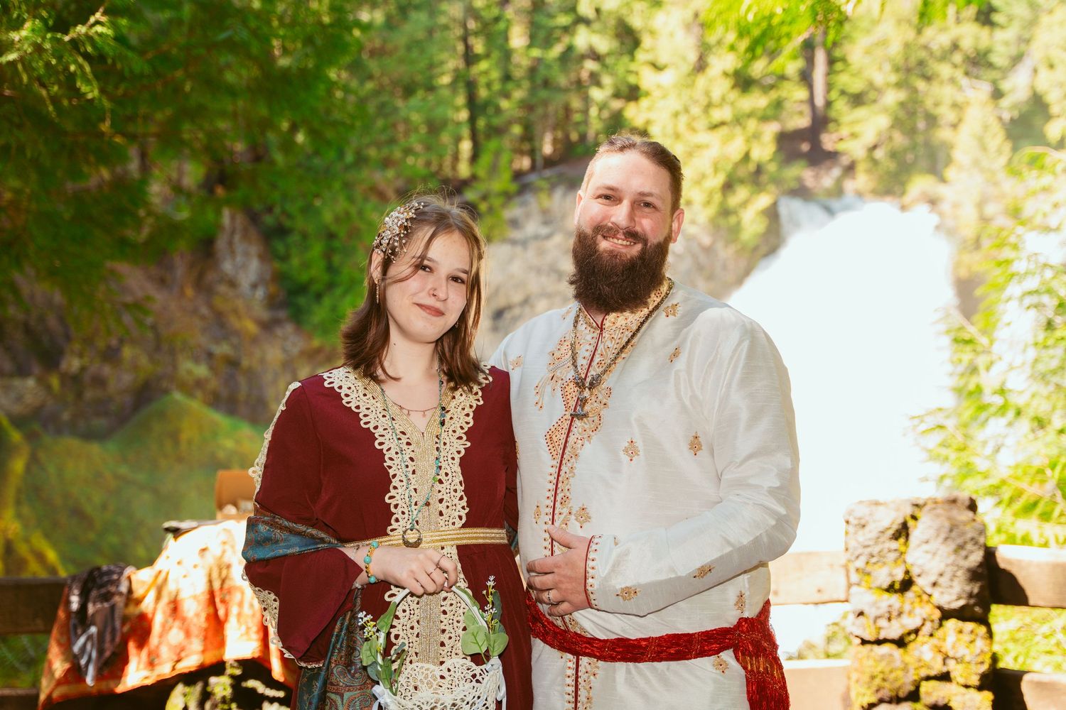 A couple poses in medieval-inspired wedding outfits with burgundy and cream colors against a forest backdrop.