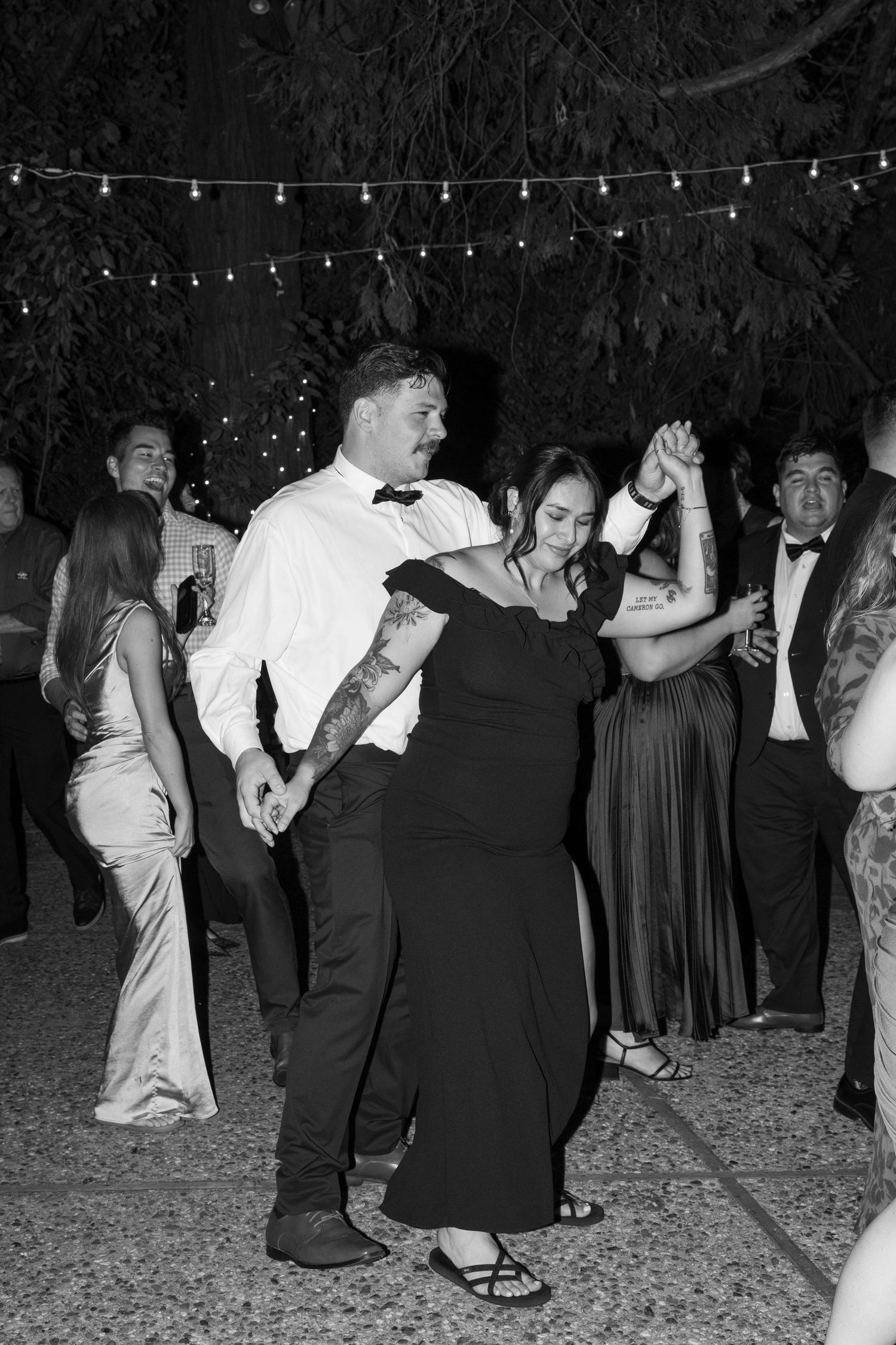 Black and white photo of couples dancing at a nighttime wedding reception under string lights.