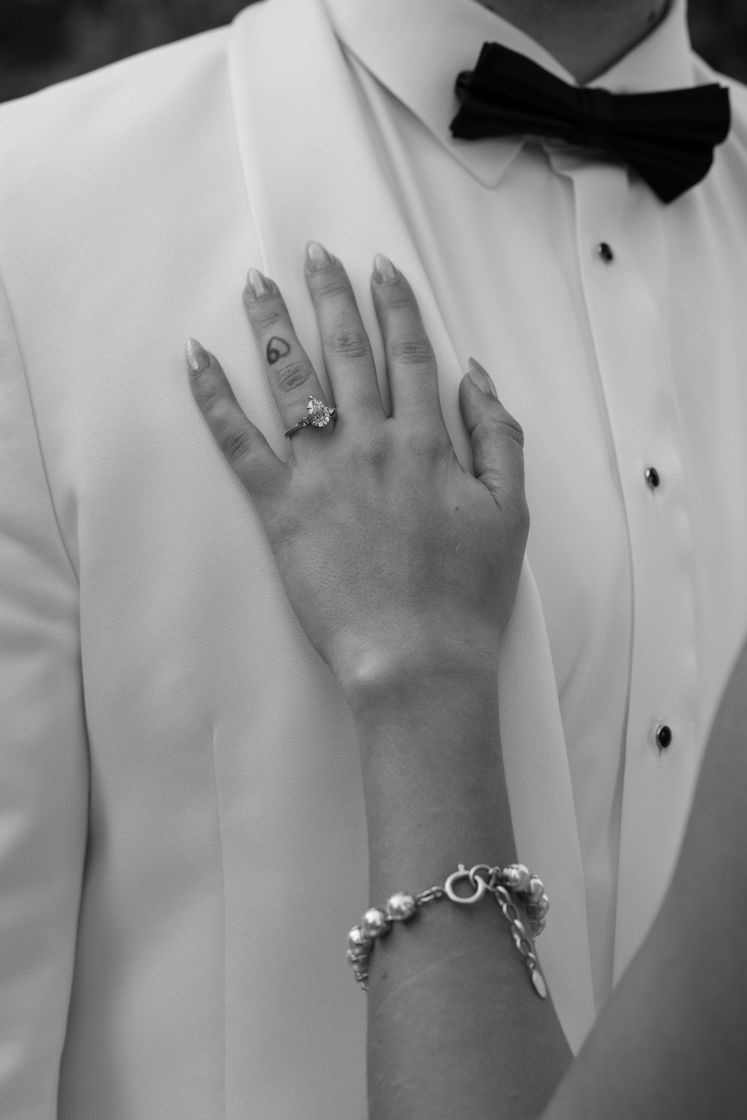 Close-up black and white shots of hands against a white tuxedo jacket showing wedding rings and jewelry.