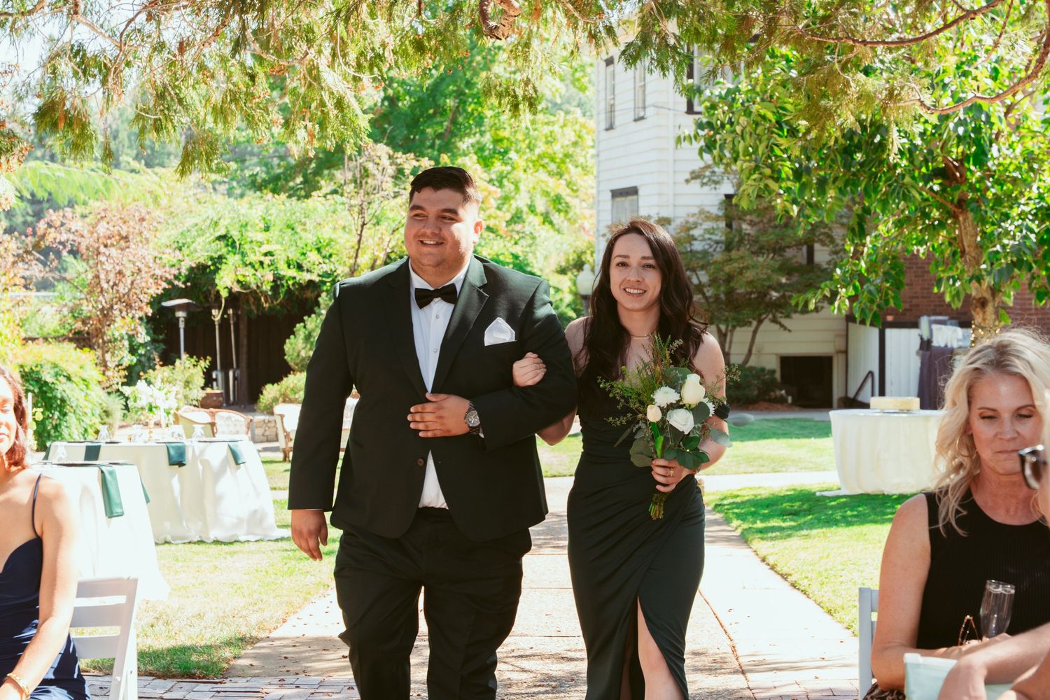 Wedding party members walk together outdoors during a sunny garden ceremony with table settings visible in background.