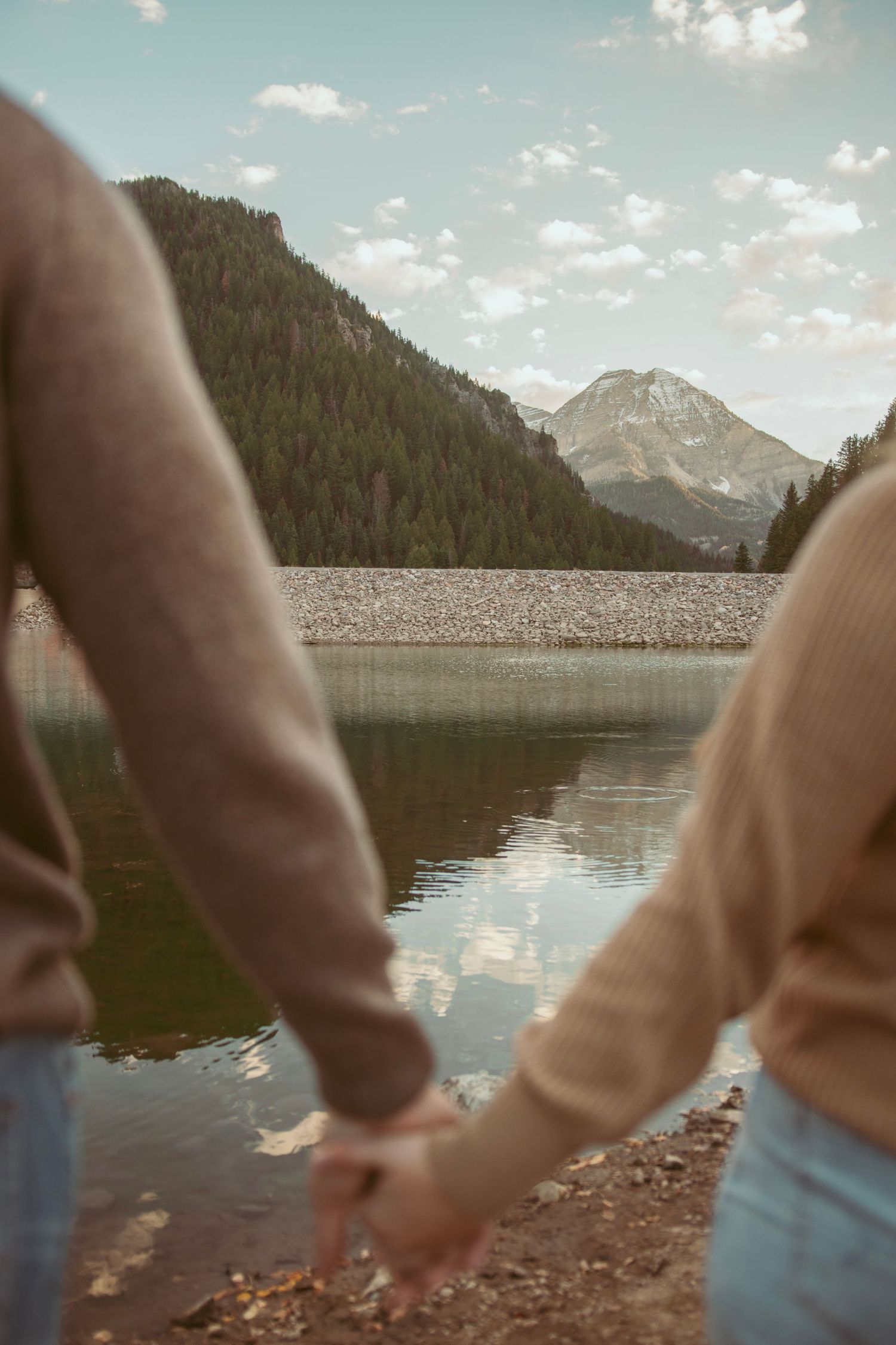 A couple holding hands by a serene mountain lake with evergreen forests and snow-capped peaks in the background.