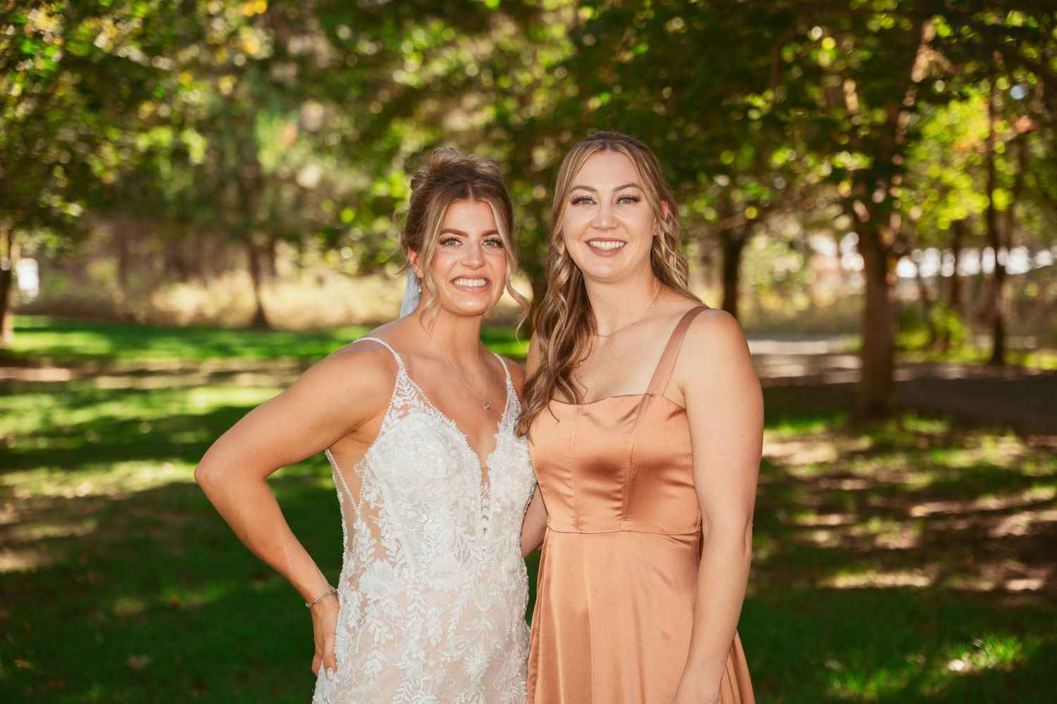 Two friends pose together outdoors in formal attire with a white lace gown and peach dress against a leafy backdrop.