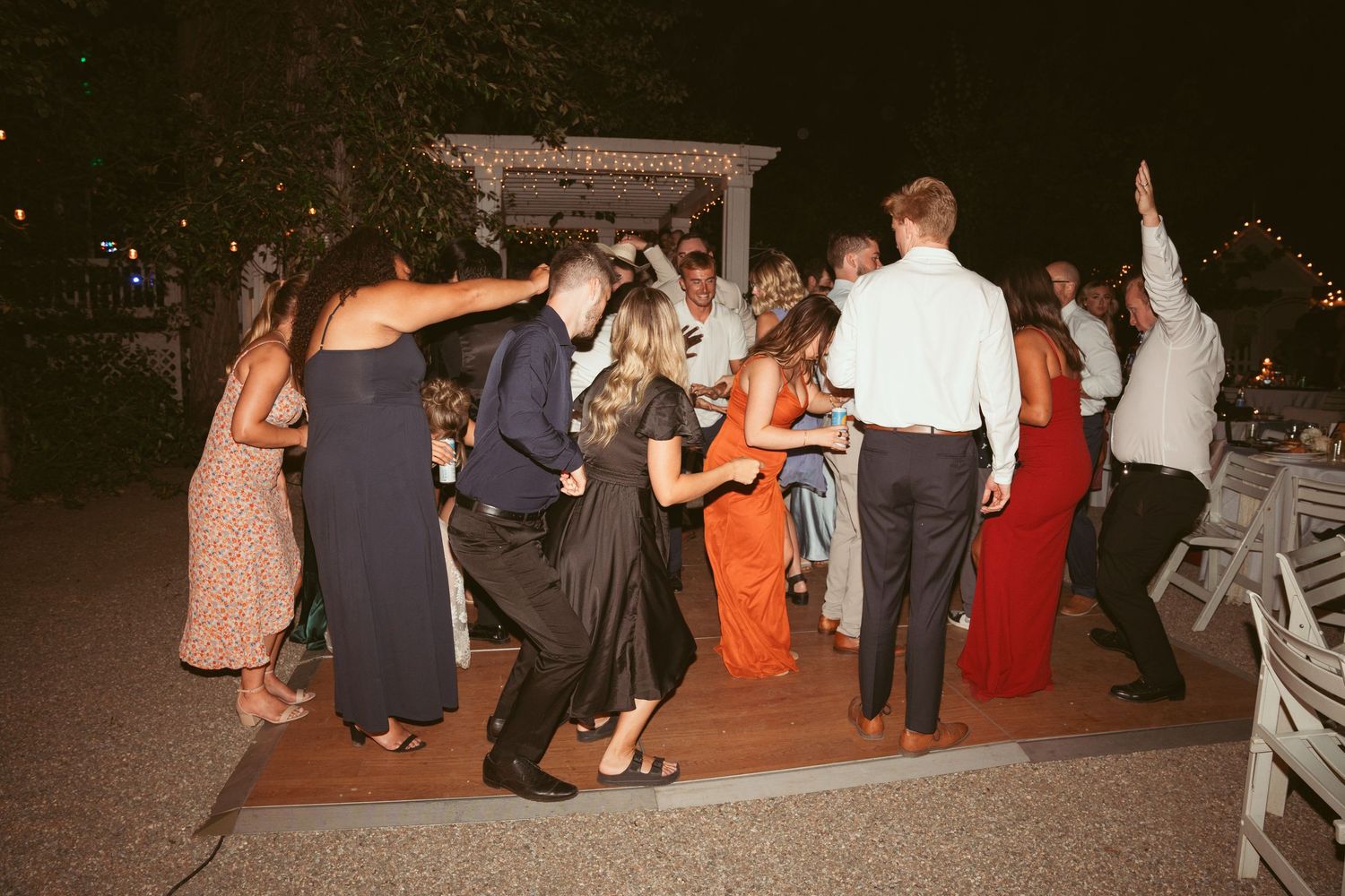 Wedding guests dance together on a wooden dance floor at an evening reception celebration.
