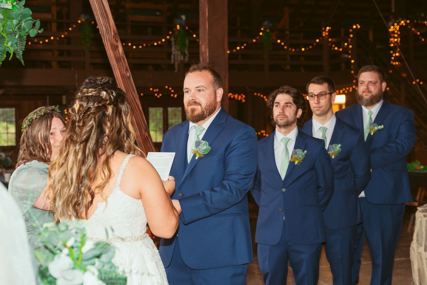Wedding party in navy suits and sage dresses standing in rustic barn venue.