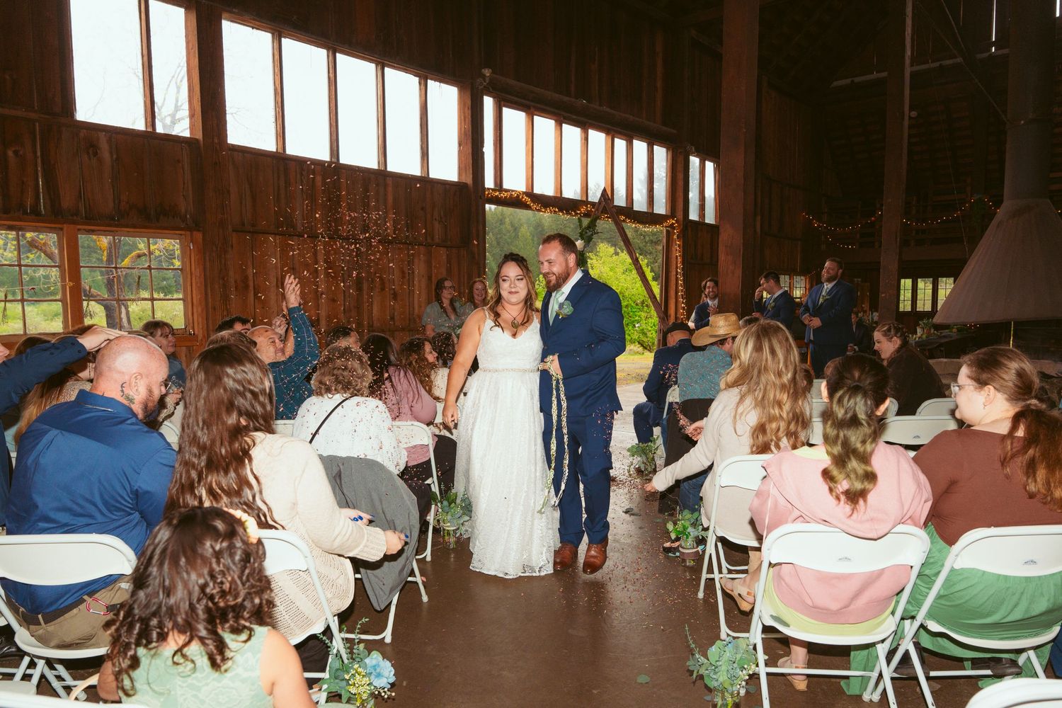 Wedding attendees seated in white chairs during an indoor barn ceremony.
