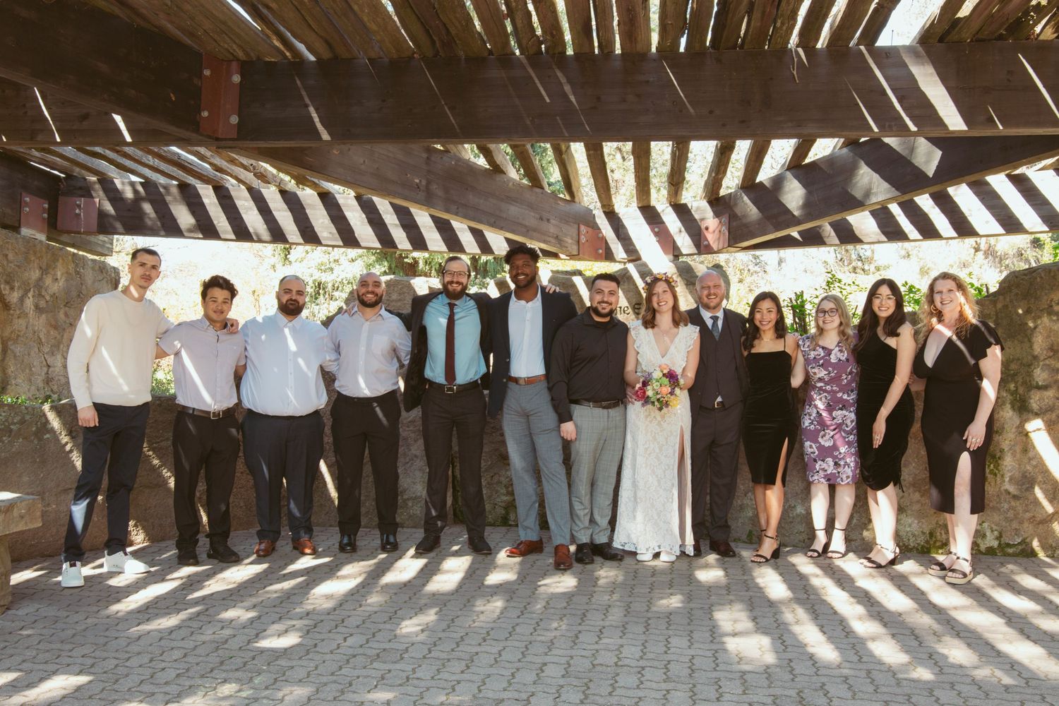 Wedding party poses together under a wooden pergola for a group photo.