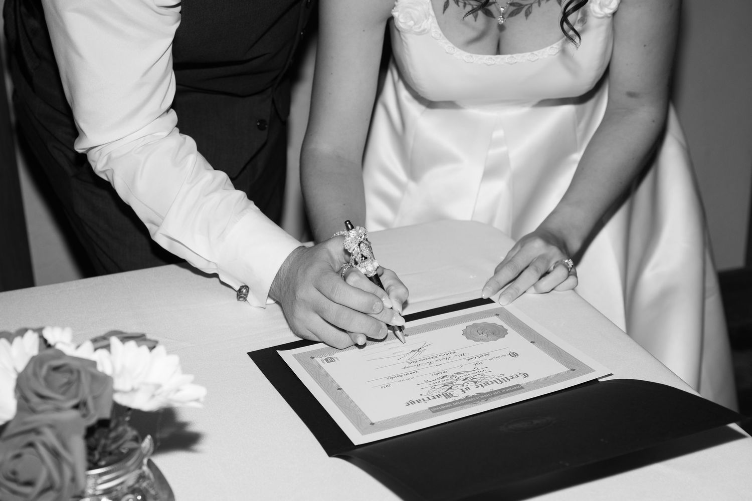 Black and white photo of hands signing a marriage certificate with roses in the foreground.