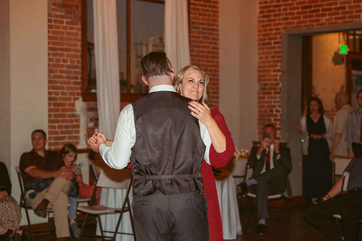 Couple dancing together in a rustic brick venue with string lights and white curtains.
