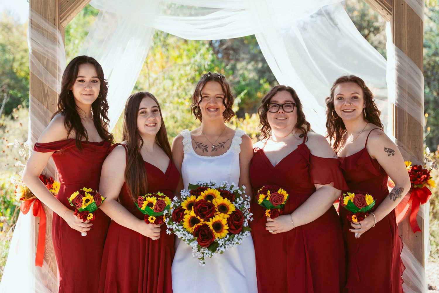 Bridesmaids in burgundy dresses holding sunflower bouquets pose under decorated pavilion.