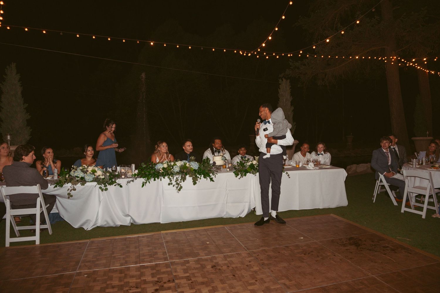 Wedding guests dance under string lights at an outdoor evening reception celebration.