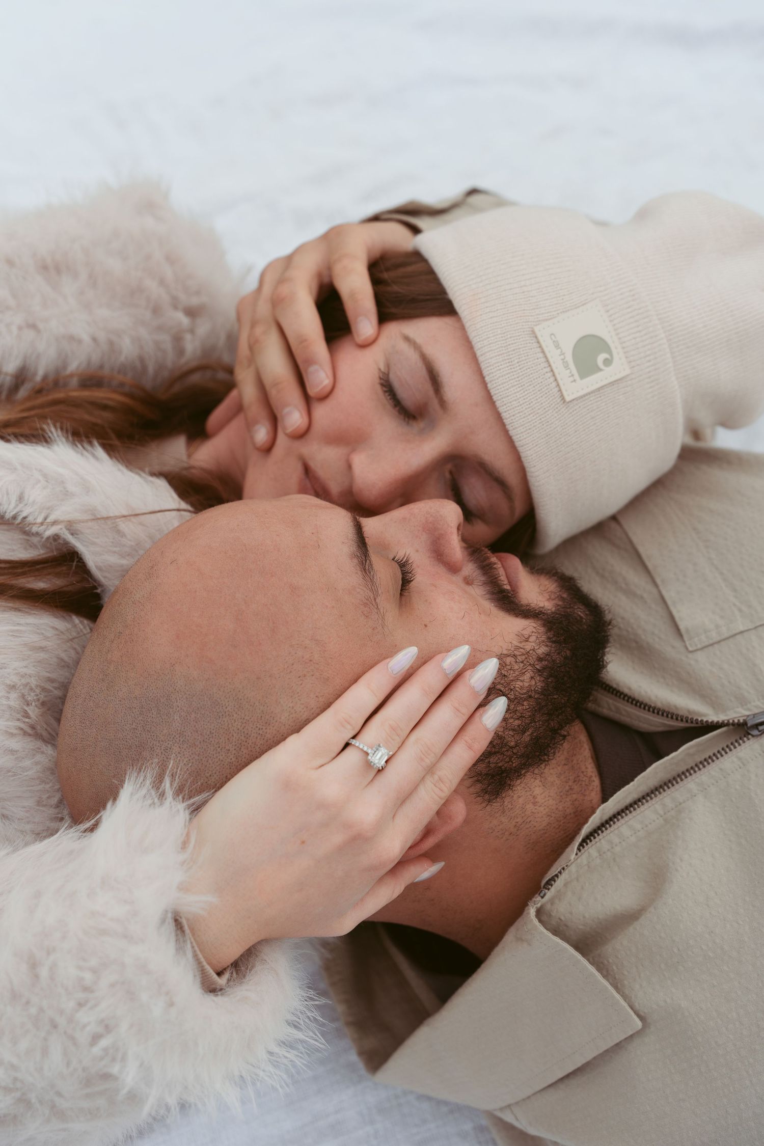 A couple lies close together in soft lighting wearing neutral colored clothing.