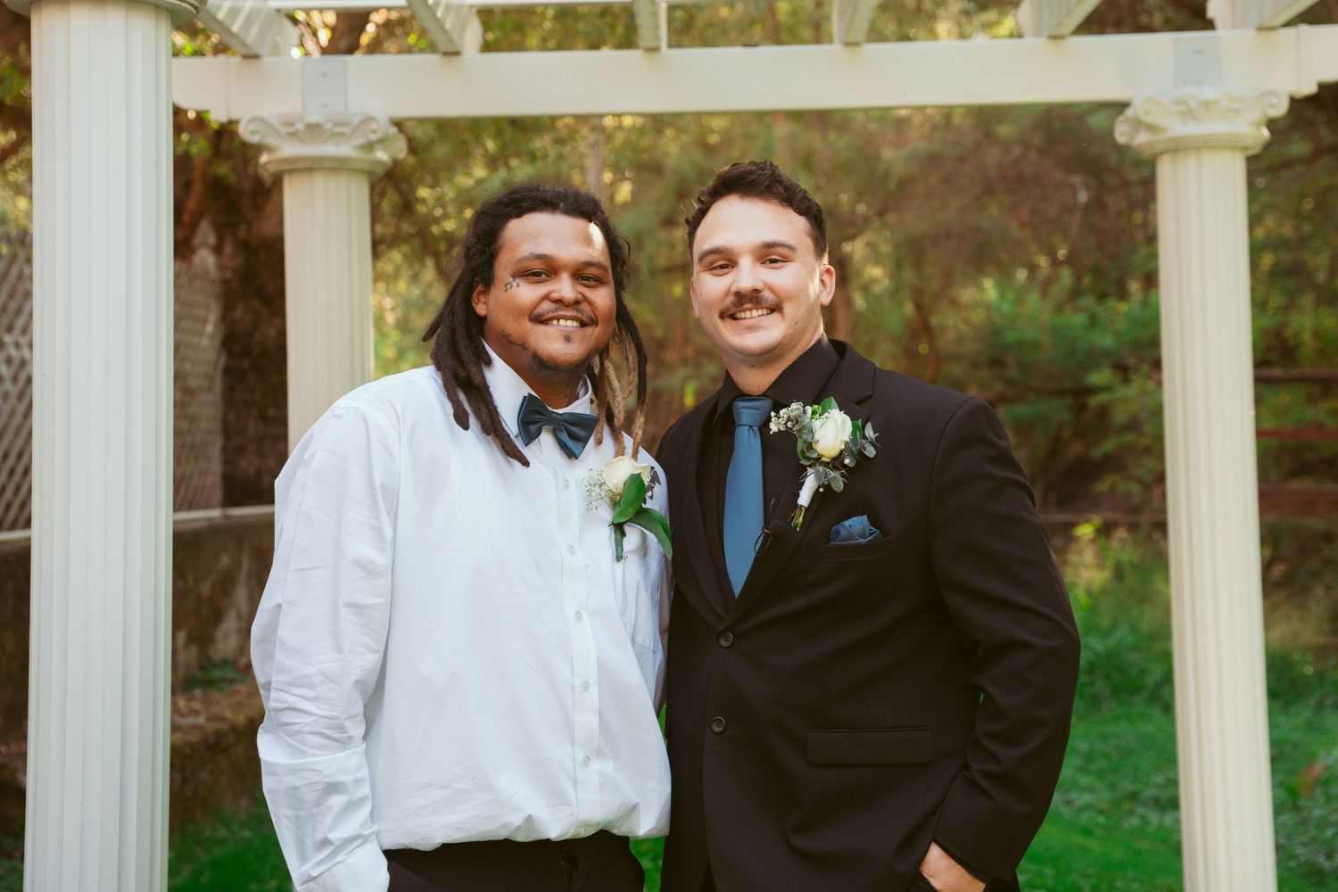 Groomsmen in formal attire share friendly moment under garden pergola.