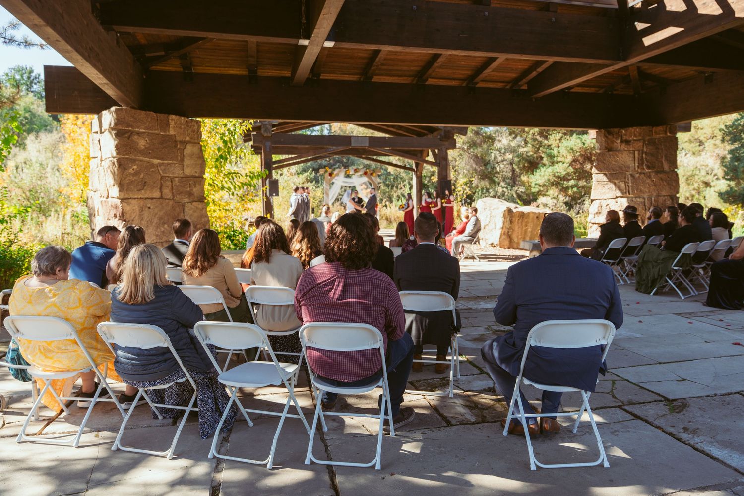 Wedding guests seated in folding chairs under a covered outdoor pavilion during a ceremony.