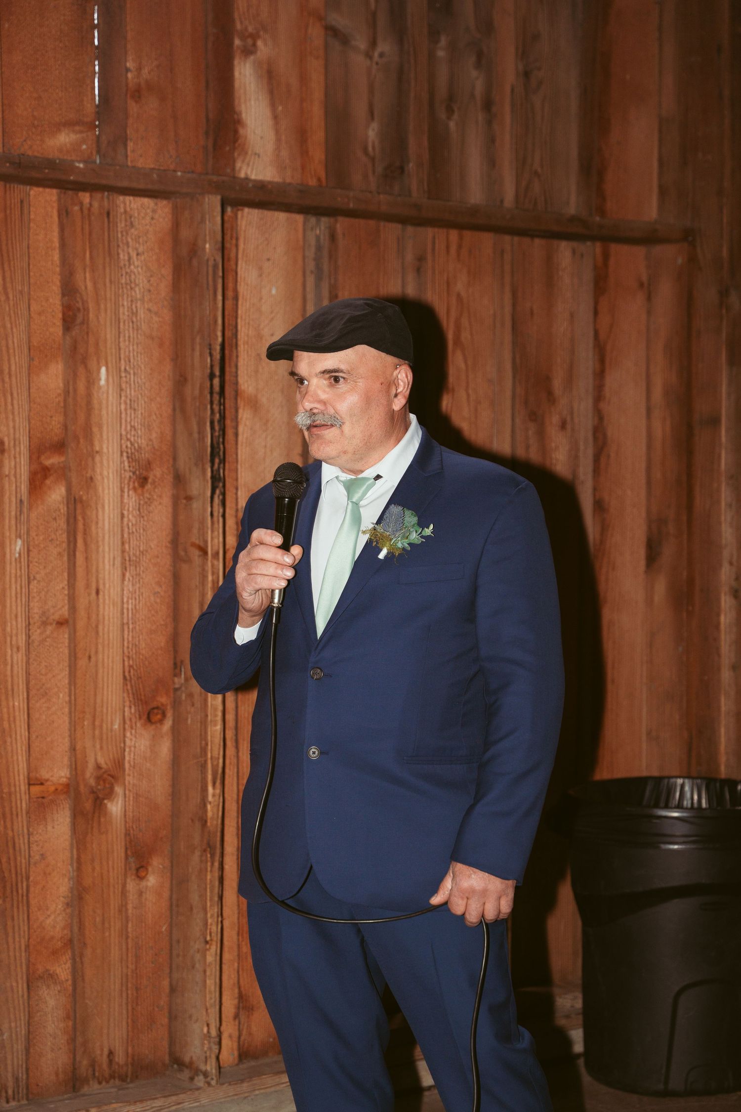 Groomsman in navy suit and cap giving animated speech at barn wedding.