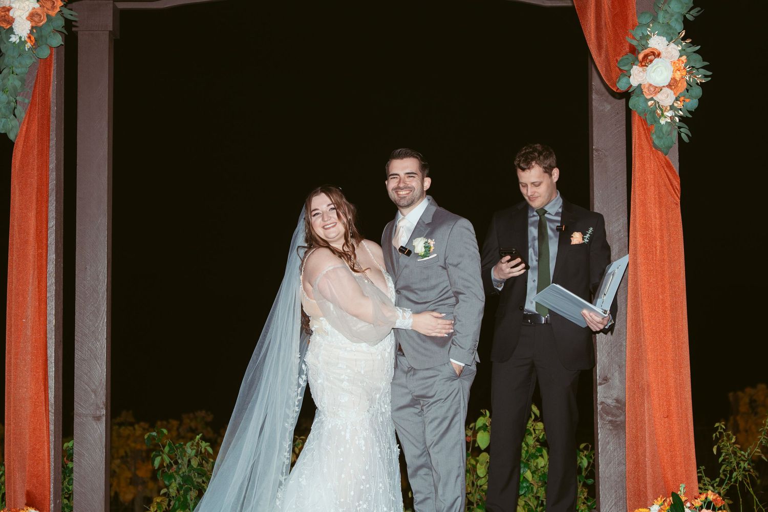Wedding party gathered under a decorated arch during an evening outdoor ceremony.