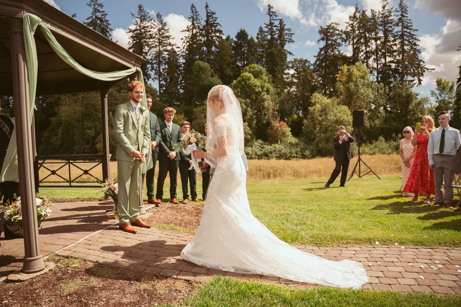 Outdoor wedding ceremony under a wooden pavilion with evergreen trees in the background.