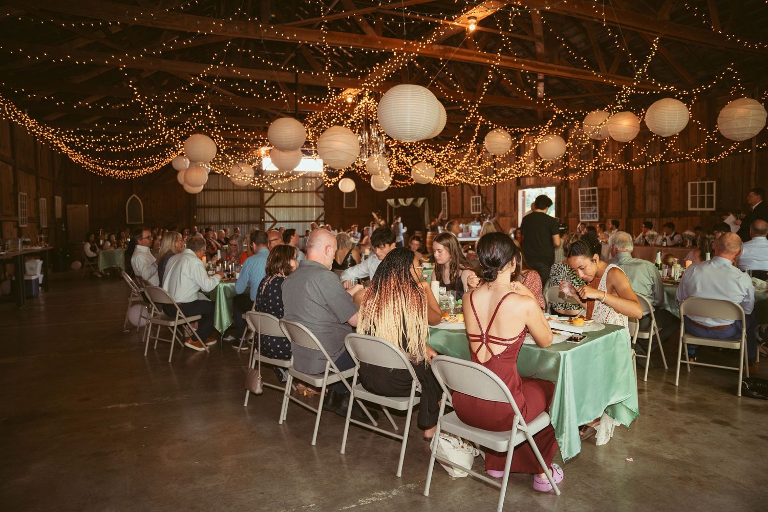 Wedding guests seated at round tables under decorative string lights and paper lanterns in a rustic barn venue.