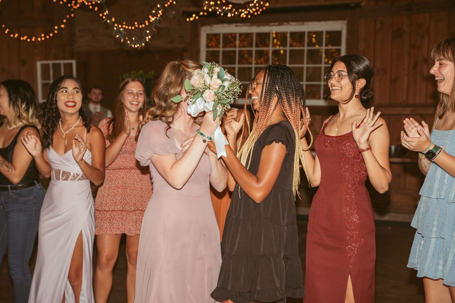 Wedding guests in pastel dresses dancing together under warm string lights at a barn reception.