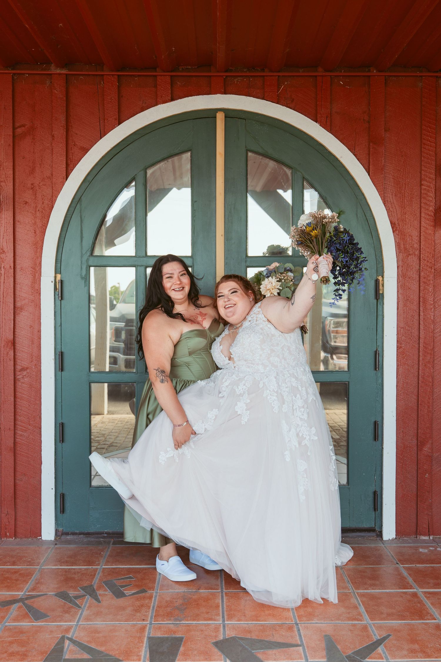 Bride in white dress lounges gracefully against arched doorway of red barn with teal trim.