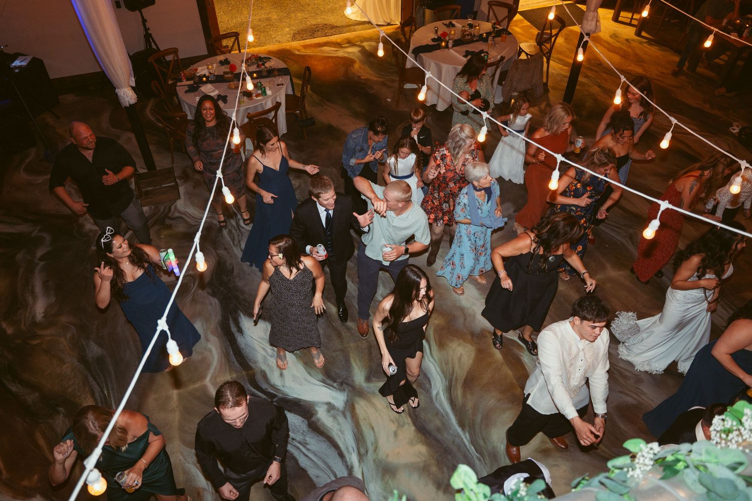 Overhead view of wedding guests dancing together under string lights at reception.