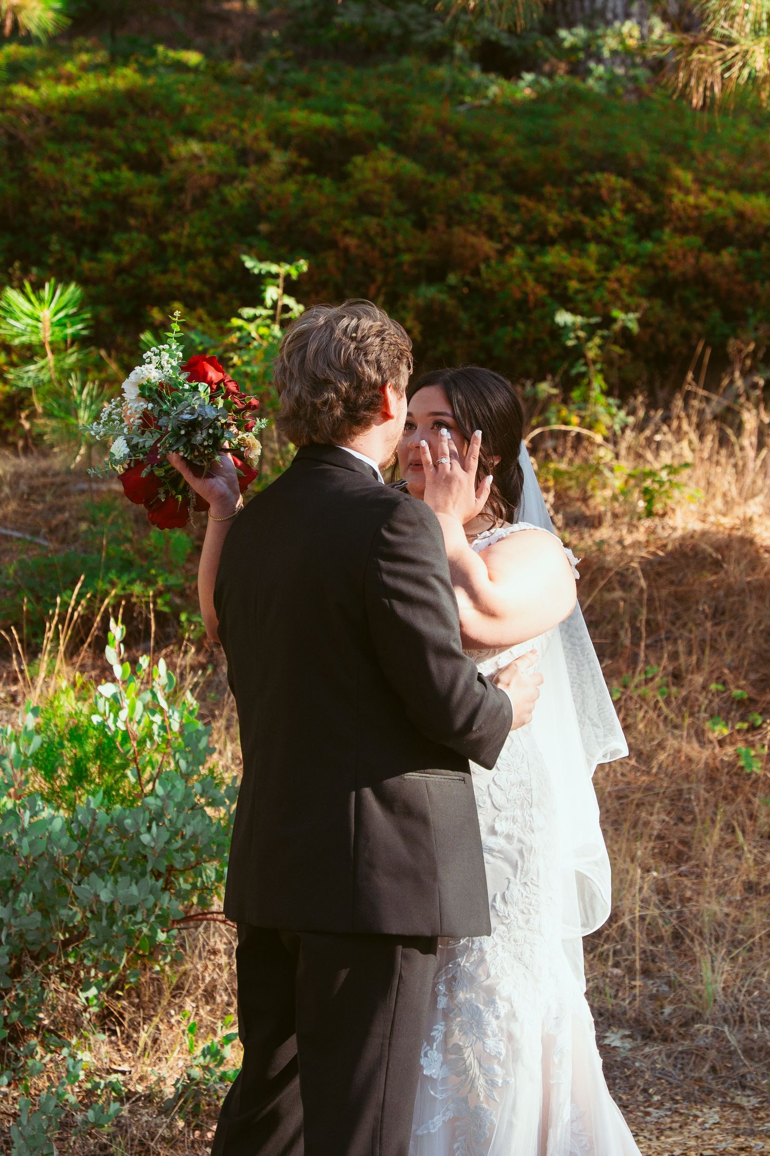 A romantic outdoor wedding moment captured in a sunlit forest setting with pine trees in the background.