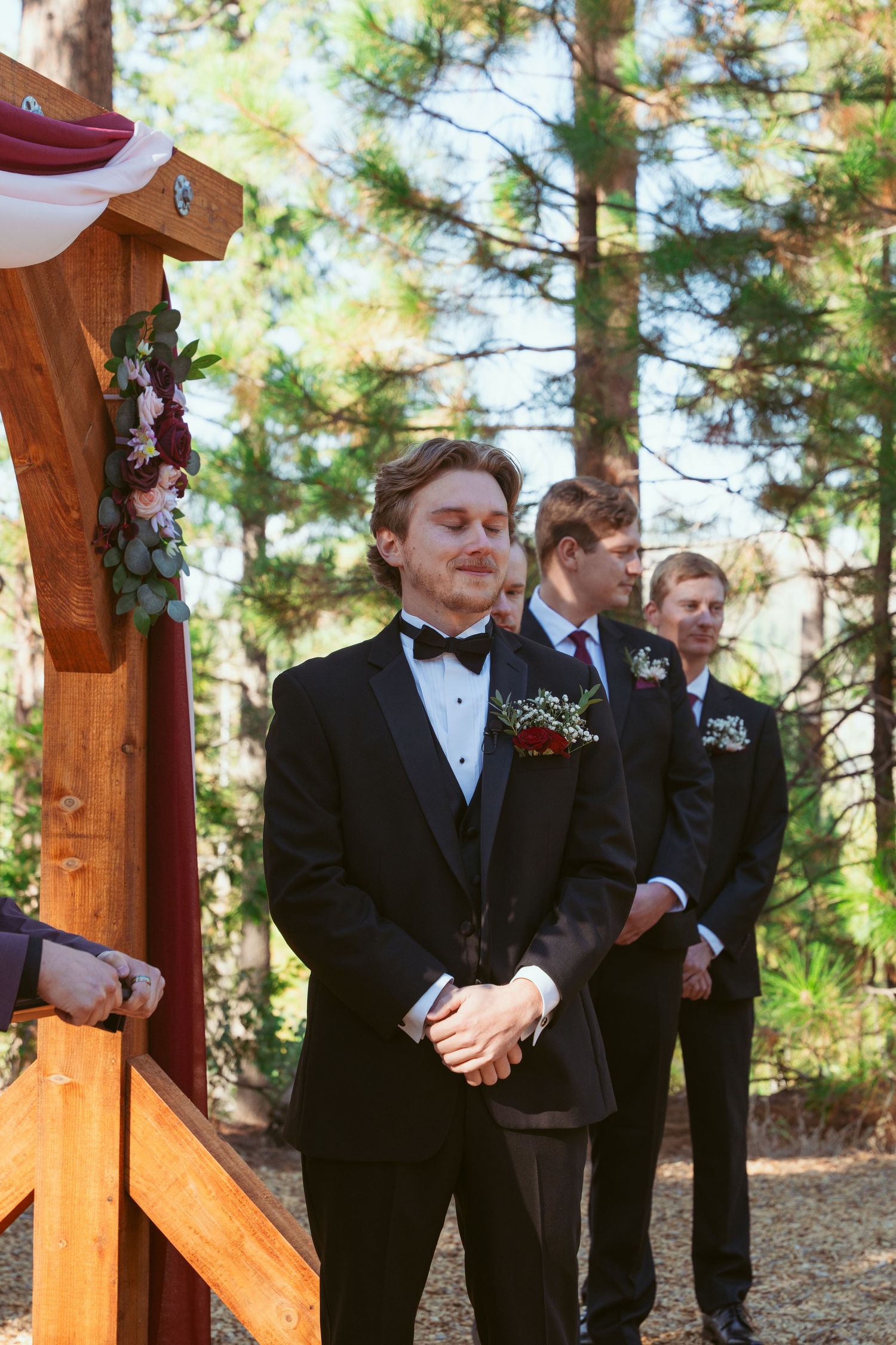 Groomsmen in black tuxedos stand at decorated wooden wedding arch in forest setting.