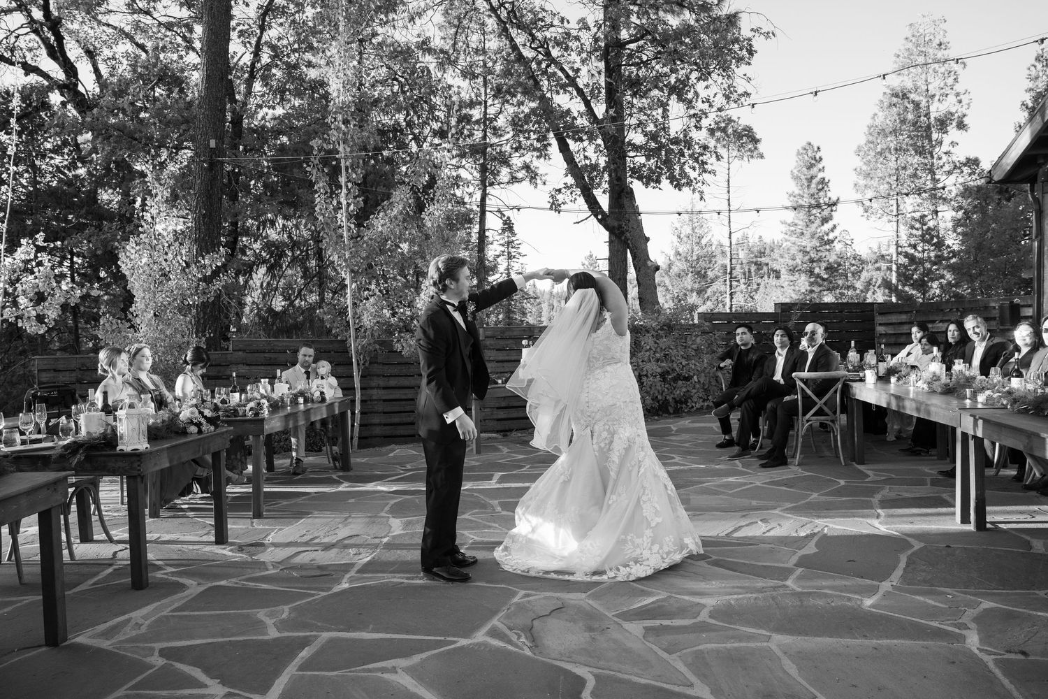 Black and white photo of newlyweds dancing on outdoor stone patio with guests watching from wooden benches.
